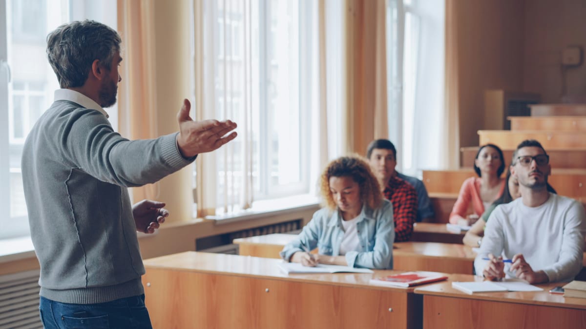 Un profesor gesticula mientras estudiantes adultos, sentados en sus escritorios, escuchan y toman notas.